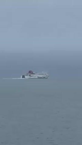 The video shows a Stena Line ferry traveling on a body of water under an overcast sky. The ferry is white with a red top and the words "Stena Line" printed on the side.  It is moving away from the camera, leaving a white wake behind it.  The water is a calm gray color. The sky is a uniform gray, suggesting fog or heavy cloud cover.