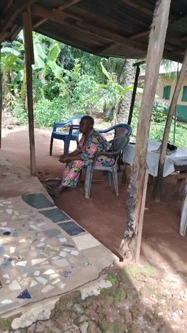 An outdoor video clip filmed in Akwa Etiti, Nigeria, shows an elderly woman sitting calmly on a dark plastic chair. She is wearing a vibrant, floral-patterned dress or caftan. In front of her is a small, raised concrete platform adorned with mosaic tiles in various shades of grey, blue, and white. The ground surrounding the platform and under the sheltered area is dirt.

The woman is positioned within an open-sided structure, likely a porch or a simple shelter, featuring a wooden frame and a corrugated roof that casts shadows on the ground. Another blue plastic chair is visible to her left. To her right, partially obscured, is a wooden table covered with a white cloth.

The background is filled with lush green tropical vegetation, including banana plants and other trees, indicating a rural or semi-rural setting. Sunlight suggests it is daytime, possibly late morning or early afternoon. No specific activities are occurring other than the woman sitting. No visible text is present in the video.