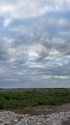 The video shows a static view of a cloudy sky over a grassy field with debris in the foreground. The sky is filled with gray and white clouds, some of which are illuminated by sunlight. In the distance, a line of trees and buildings can be seen. The foreground is cluttered with piles of concrete and rubble. The overall scene is one of an undeveloped or abandoned area under a dramatic sky.