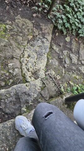 The video depicts a person's legs and feet walking down a stone path in a natural setting. The person is wearing ripped grey jeans and white sneakers. The path is made up of irregular stones, and there is a significant amount of greenery, including ivy and moss, growing along the sides of the path and between the stones. A metal handrail can be seen on the left side of the path, suggesting the path is a designated walking trail. The perspective is from the person's point of view as they walk, giving the viewer the sense of being on the trail with them. Towards the end of the video, other people's legs can be seen ahead on the trail.