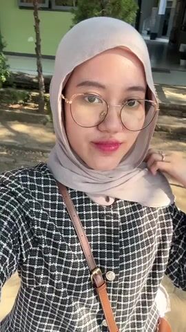 A young woman wearing a beige hijab and glasses smiles and poses for a selfie outdoors. She is wearing a black and white checkered jacket and a brown purse is slung across her body. In the background, there are trees and a building. The woman adjusts her hijab and flashes her teeth, revealing braces.