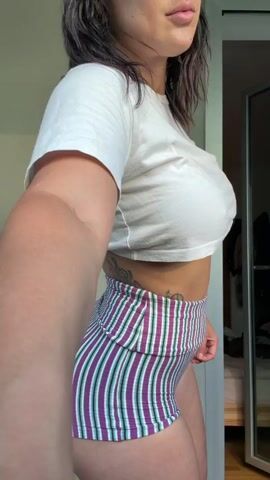 A woman is captured in a vertical video, likely indoors. She wears a white short-sleeved crop top and high-waisted, form-fitting shorts with purple, green, and white vertical stripes. Tattoos, including a musical note, are visible on her left side/ribs.

The video shows her initially posing, flexing her right arm. She then puts on a white baseball cap and dark sunglasses, smiling as she does so. She continues to move her body and arms, seemingly dancing or posing to music, maintaining a side profile to the camera. The background is a plain light-colored wall with a reflective surface, possibly a wardrobe or mirror, visible on the right. The lighting appears bright, suggesting daytime. No visible text is present in the scene.