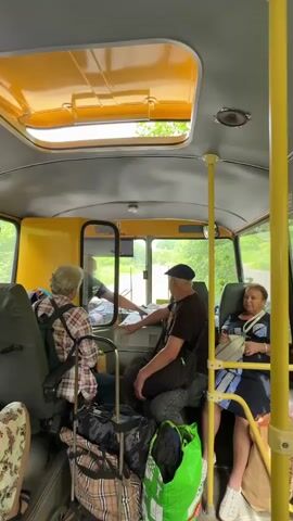The video shows the interior of a yellow bus. In the foreground, a woman with gray hair and wearing a plaid shirt is seated in the driver's seat, facing forward. To her right, a man wearing a dark t-shirt and a blue cap is seated, looking forward. He has his hand on a panel. To the right of the man, a woman with gray hair wearing a blue and white patterned dress is seated, looking forward. Luggage is visible in the foreground and between the seats. A sunroof is open above the driver. The scene outside the bus appears to be a rural road with trees and greenery. The driver is handing a piece of paper, possibly a ticket or fare, to the man.