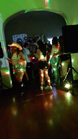 The video captures a 20th birthday party in an indoor setting. The room, which features an arched entryway, is dimly lit with colorful party lights creating a dynamic ambiance.  Several young people can be seen, including some in cowboy hats. One woman with a white crop top and short black skirt walks towards the camera holding her cell phone. Behind her, a group of people are mingling near a table decorated with balloons spelling out "20". A pair of speakers on stands flank the crowd, and a man can be seen hugging a woman. The room is well-lit.