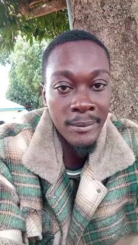 The video is a selfie-style recording of a dark-skinned man with short, dark hair, a mustache, and a goatee. He is wearing a plaid jacket with green and brown checks and a fluffy cream-colored collar. In the background, there is a large tree trunk to his right, and greenery and some objects that appear to be buckets or containers on the left. The man looks directly at the camera at times, and then glances away, seemingly engaged in conversation or observing something off-camera. His facial expressions change from a slight smile to a more neutral or questioning look. The lighting suggests it is daytime and outdoors.