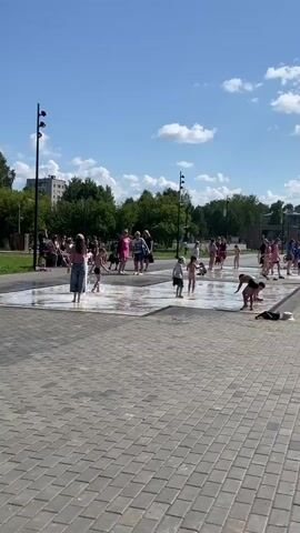This is a short video clip of a splash pad on a sunny day. Several children and some adults are visible in and around the splash pad. The splash pad has water jets spraying upwards from the ground, creating a shallow pool. Some children are running through the water, while others are crouching or sitting in it. Adults are standing around the perimeter, watching the children. The background shows trees, buildings, and a blue sky with white clouds. The ground around the splash pad is paved with interlocking stones. The overall scene depicts a recreational activity on a warm day.