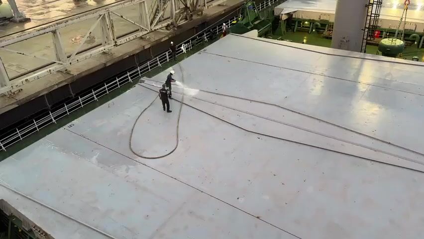 Two individuals wearing hard hats and dark work clothing are on the deck of a ship. One individual is holding a hose and spraying water onto the deck. The other individual is standing next to them, also appearing to be involved in the cleaning process. A coiled hose lies on the deck between the two people. The deck is a large, flat, gray surface with several long, thin, raised lines running across it. In the background, there is a raised walkway or structure with metal railings and industrial equipment. The environment appears to be an outdoor, industrial setting, likely a port or shipyard.