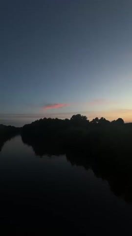 The video shows a wide shot of a natural landscape at dusk. The sky is a gradient of deep blue to pale orange, with thin clouds. A dark silhouette of trees forms a horizon line across the middle of the frame. In the foreground, a body of dark water reflects the trees and sky. The overall impression is one of quiet and stillness.