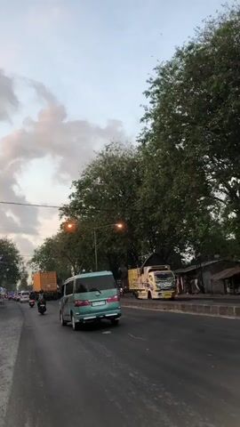 The video shows a street scene in Kejapanan, Indonesia. The road is paved and appears to be a busy thoroughfare, with several vehicles present. In the foreground, a teal-colored van is driving away from the camera, its brake lights illuminated. To the left of the van, a motorcycle carrying a rider is also moving forward. Further down the road, a large yellow truck is visible, its trailer laden with cargo. On the right side of the road, a colorful truck with intricate designs is parked. Several other motorcycles are parked along the side of the road, and a few people can be seen in the background, suggesting a commercial or residential area.

The scene is set during what appears to be either dusk or dawn, indicated by the soft, diffused lighting and the cloudy sky overhead. The trees lining the road are full and green, suggesting a tropical or subtropical climate. Streetlights are visible, casting an orange glow, and are illuminated, further supporting the idea of low light conditions. The overall atmosphere of the scene is one of everyday life and transit in a populated area. No discernible text is visible in the video.