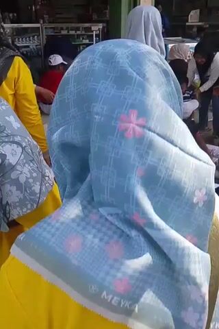 The video shows a group of people, mostly children and adults wearing hijabs, gathered outdoors. Some children are sitting on the ground, and one child has a blue bib with the number 81 on it. There are some food items and drinks on the ground. In the background, there are shops or stalls with shelves stocked with various items. A motorcycle is parked to the right of the frame. The overall atmosphere appears to be informal, possibly a gathering or an event. The lighting suggests it is daytime, with sunlight filtering through.