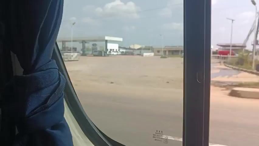 The video, taken from inside a moving vehicle, shows a view of a gas station through a window. A blue curtain partially covers the window. The gas station has multiple fuel pumps under a large canopy. There is green grass between the road and the gas station.  In the background, there are other buildings.  The camera then shifts to show the interior of the vehicle, including the headrest of a seat and a person with curly hair sitting in front.  The overall atmosphere is bright and somewhat hazy due to the weather conditions.