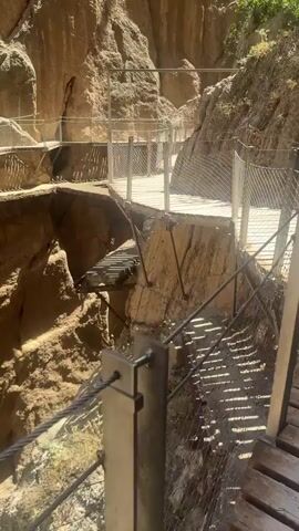 The video shows a narrow pathway constructed along the side of a rocky canyon. The pathway is made of wooden planks with metal railings and netting for safety. The canyon walls are steep and made of rough, brown rock. Sunlight casts shadows on the pathway. The pathway appears to be elevated above the canyon floor, with a steep drop on one side. The video pans to the right, showing the pathway continuing along the canyon wall, with vegetation growing on the canyon rim. The scene appears to be a tourist attraction or hiking trail.