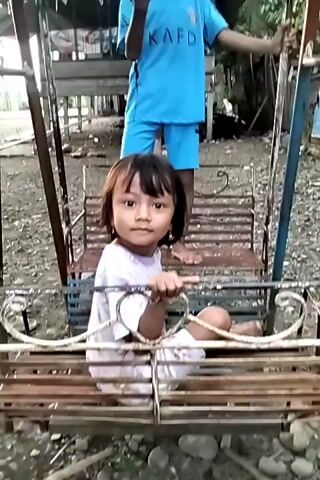A low-angle shot depicts two barefoot children on a rusty swing set in an outdoor setting. A young girl with bangs, wearing a white shirt with a pattern, sits on the swing, holding onto the bar. Behind her, a boy in a matching light blue shirt and shorts set stands on the swing seat, facing the camera, with his hands near his mouth. 

The swing set is made of metal with peeling paint. The ground beneath the swing set appears to be dirt and small rocks. In the background, a wooden structure with a corrugated metal roof is visible, surrounded by greenery and other undefined objects. The lighting suggests that the video was taken in the late afternoon or early morning.