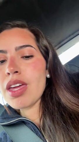 The video shows a woman speaking, while seated in a car and wearing a seatbelt. She has long brown hair, brown eyes, and is wearing makeup, including blush and lipstick. She also has pearl earrings and a gold necklace. The camera angle is a close-up, showing her face from a slightly low angle. The interior of the car is visible in the background, including the car’s roof and side window.