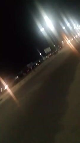 A dimly lit scene shows a street at night. The viewpoint is from the edge of the road, capturing the asphalt surface with faint details. A line of parked cars extends into the distance on the left side of the road. Streetlights cast bright, streaky beams across the scene, obscuring details.  Additional lights, possibly from moving vehicles, are also visible in the distance, contributing to the overall brightened yet hazy effect. The right side of the frame is dark, and the top portion shows a dark, featureless sky.