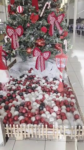 The video shows a decorated Christmas tree in an indoor shopping center. The tree is green and decorated with red and white striped bows, red and white lollipop decorations, candy canes, and shiny red ornaments. The tree stands on a base covered with a layer of white material that resembles snow. Surrounding the base is a collection of white, red, and dark gray ornaments contained by a short white picket fence. A red and white candy cane-striped lamppost sits on the base beside the tree. In the background, there are stores and people walking through the shopping center. The lighting is bright, typical of a commercial space.