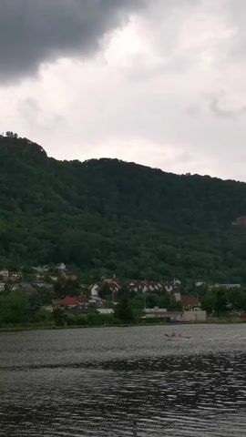 A view from a body of water looking towards a village nestled at the base of a densely forested hill under a cloudy sky. In the distance, a small boat with at least two people rowing is visible on the water. The village consists of houses with red roofs, and a few scattered trees. The hill is covered in dark green foliage. The sky is overcast with grey clouds.