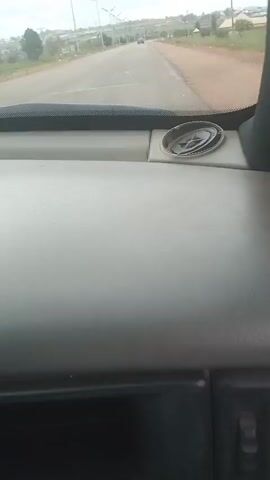 The video captures a view from inside a vehicle, looking out through the windshield. The dashboard is visible in the foreground, with a light gray, textured surface. A circular, decorative object, possibly a compass or emblem, is placed on the dashboard to the right. Beyond the dashboard, a paved road stretches into the distance, bordered by vegetation and some buildings on either side. The sky is overcast. Several vehicles, including what appears to be a car, are visible on the road ahead. The overall scene suggests the vehicle is in motion on a rural or suburban road.