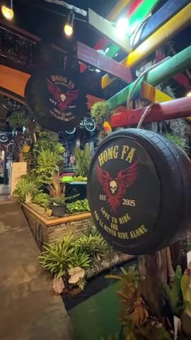 The video shows the exterior and interior of a establishment, likely a restaurant or bar, with a motorcycle theme. The exterior features a large, circular sign with the text "HONG FA EST 2005 LOVE TO RIDE YOU'LL NEVER RIDE ALONE". There are also many plants surrounding the entrance. Inside, the space is decorated with motorcycle tires, various pictures and art, and at least one bicycle is hanging from the ceiling. Wooden tables and chairs are set up for customers. A person is seen walking away from the camera into the establishment. There is also a sign that says "OPEN" in both Thai and English. The lighting suggests it is nighttime. The overall atmosphere appears to be casual and welcoming, fitting for a biker-friendly establishment.