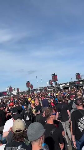 A wide shot shows a large crowd of people standing and milling around outdoors under a cloudy sky. Many people are wearing black shirts and hats. In the background, there are large screens and a stage setup for a concert or event. Some people are holding drinks and food. The ground appears to be asphalt or pavement.