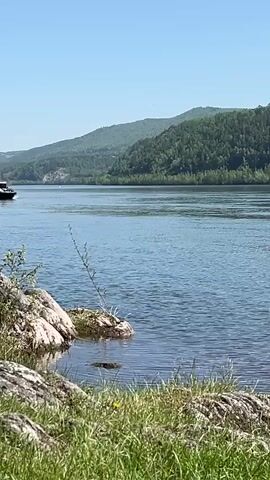 A large, dark-colored hovercraft travels rapidly across a wide, calm body of water from left to right. The vessel kicks up a substantial spray or wake as it moves. In the foreground, a rocky shoreline with patches of green grass frames the bottom of the shot. The background features a distant, undulating mountain range densely covered in green trees, indicating a forested landscape. The sky is clear and bright blue, suggesting a sunny day. No people are explicitly visible. The scene depicts a serene natural environment contrasted with the powerful, fast-moving hovercraft. There is no visible text. The setting appears to be a river or large lake, consistent with the provided location context of Sliznevo, Russia.