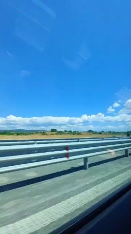 This is a first-person perspective video taken from inside a vehicle, likely a bus or car, moving along a highway. The view is through the window, which has some reflections on it. The scene outside consists of a clear, bright blue sky with scattered white clouds. In the middle ground, there is a highway guardrail made of parallel metal bars. Beyond the guardrail, there is a landscape of dry, brown vegetation and green bushes or trees. The road surface appears to be asphalt. The vehicle is in motion, indicated by the slight blur and the movement of the scenery. The lighting suggests it is daytime, likely during sunny weather.