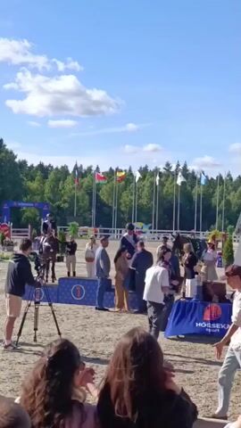 A crowd of people are gathered around a podium and a horse in an outdoor equestrian event. The scene appears to be an awards ceremony, with individuals in formal attire standing on a blue podium with a "1" visible. One person is mounted on a horse. A banner with the text "HORSESPORT" is visible on a table with trophies. The sky is blue with scattered clouds, suggesting a pleasant day. Trees form a backdrop to the event. The general atmosphere appears to be one of celebration and achievement. The time of day is likely daytime given the bright lighting and clear sky.