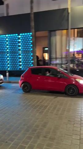The video shows a red car driving down a street at night. There are people walking on the sidewalk and some businesses with lights on. The street is paved with cobblestones. The car is moving from left to right across the frame. The traffic light is red, and people are waiting to cross the street. There are also streetlights and building lights illuminating the scene.