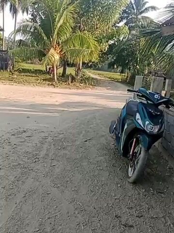 A dark teal and black scooter is parked on a dirt road next to a weathered wooden building. Palm trees and other foliage are visible in the background under a bright sky with scattered clouds. The scooter is angled towards the camera and is the primary focus of the shot. The building appears to be a small structure, possibly a shed or outhouse, with a corrugated metal roof. The road is dusty and uneven. There are no people visible in the frame. The lighting suggests it is daytime, likely late afternoon or early morning due to the warm tones.