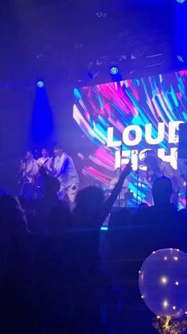 A dimly lit indoor venue with a stage and a large screen displaying colorful abstract patterns. On stage, a band is performing. A male singer in the center, wearing light-colored clothing, is holding a microphone and singing. Other band members, also in light-colored attire, play instruments such as guitars and what appears to be an accordion. The audience in the foreground is silhouetted, with some individuals raising their hands in the air. One person in the foreground is holding a balloon with lights inside. The overall atmosphere suggests a live musical performance.