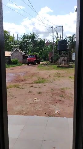 The scene is viewed through an open doorway. Outside is a dirt lot, with patchy grass and scattered trash. A red truck, filled with green foliage, is positioned on the dirt road in the lot. Behind the truck, a small, weathered building is visible to the left. To the right of the truck, there is a water tower structure, along with lush greenery and palm trees. Overhead, power lines stretch across a bright, partially cloudy sky. The flooring inside the doorway is covered with light-colored square tiles.