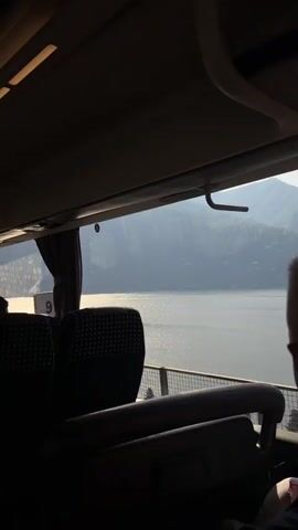 The video shows a view from inside a moving bus. The seats are black with a repeating dot pattern. A person's head and upper back are visible in the foreground. Outside the window, a large body of water is visible, with mountains in the background. The light is bright, and the water is shimmering. Curtains are partially drawn on the window. The bus is moving forward.