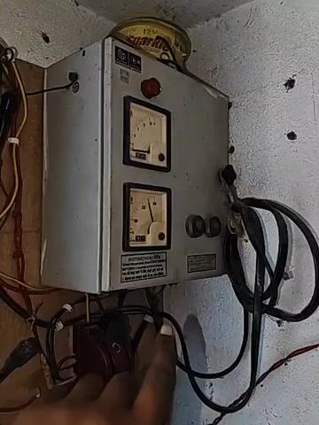 A hand turns a knob on a metal control panel. The panel has two analog meters, one labeled A and the other V, and a red indicator light that illuminates. Two black electrical cords are attached to the right side of the panel. A yellow tin can labeled "Sparkle" sits on top of the panel. The panel is mounted on a textured white wall.