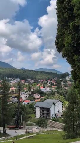 A video shows a drone view of a village nestled in a valley with mountains in the background. The drone flies over the village, showing houses, trees, and a construction site with a large drilling machine. The sky is partly cloudy with the sun shining through. The mountains are covered with lush green forests. The village appears to be a quiet and peaceful place. The video appears to be shot in the summer, as the trees are full of leaves and the weather is warm.