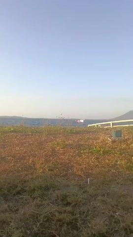 The video shows a seascape with a small boat on the water. The foreground is a grassy area with dry, brown vegetation and some green patches. There is a white fence post visible on the right side of the frame. The water is blue and appears slightly choppy, with ripples on the surface. A small boat, primarily white with some red accents, is positioned in the middle ground of the water, moving from left to right. The sky is clear and pale blue, with a hint of haze near the horizon. In the far distance, a dark landmass or mountain is visible on the right. The lighting suggests it is either early morning or late afternoon.