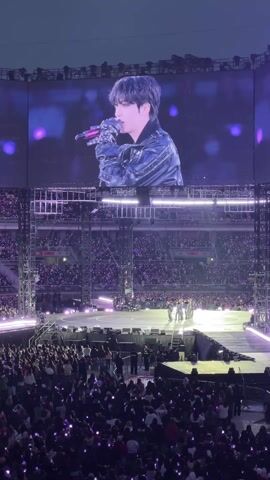The video depicts a large concert venue, likely a stadium, filled with a massive crowd. The audience members are holding up light-emitting devices, creating a sea of purple lights. On a large screen suspended above the stage, a male performer, presumably a singer, is visible. He is holding a microphone and wearing a dark jacket. Below the screen, a group of figures are gathered on a stage, possibly other members of a band or performers. The overall atmosphere suggests an exciting and energetic concert event.