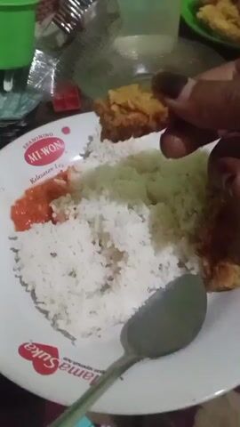 The video shows a close-up of a person's hands holding food over a plate of rice. The plate has red writing and a heart logo. There is also a spoon on the plate. The person appears to be eating. The background is blurry but suggests an indoor setting with other objects visible. The lighting is dim.