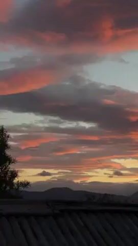 The video shows a colorful sunset with orange, pink, and gray clouds in the sky. Below the clouds, there is a view of a mountain range. In the foreground, there is a rooftop with visible seams. The camera slowly pans to the right. A building appears briefly on the right side of the screen.