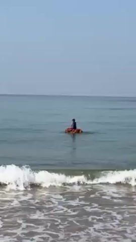 A person rows an orange raft in the ocean. The person is facing away from the viewer and wearing a blue top and dark-colored shorts. They use a double-bladed paddle to propel themselves through the water. The ocean is calm, with gentle waves approaching the shore. The sky is clear and pale blue. The sand of the beach is visible in the foreground.