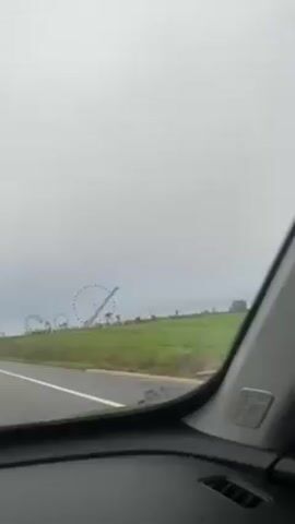 The video shows a roadside view from inside a moving vehicle. On the right side of the road, a green field features a Ferris wheel and a roller coaster. The sky is overcast, giving the scene a muted, grey tone. The road is paved and marked with white lines. The camera angle suggests the view is from the driver's seat. Other vehicles can be seen traveling on the road.