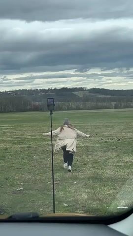 A woman runs away from the camera through a field of grass towards a distant treeline. She is wearing a beige trench coat, dark pants, and a hat. Several birds are also flying in the field. A phone mounted on a stand is visible in the lower left of the frame. The sky is overcast with dark, heavy clouds. The view is captured from inside a vehicle, with the dashboard visible in the bottom of the frame.