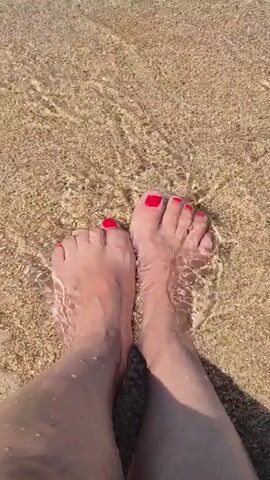 A first-person perspective video shows a pair of feet with bright red toenail polish. The feet are standing in shallow, clear water that is lapping over a sandy beach. The sunlight creates rippling patterns on the water and the sand beneath.