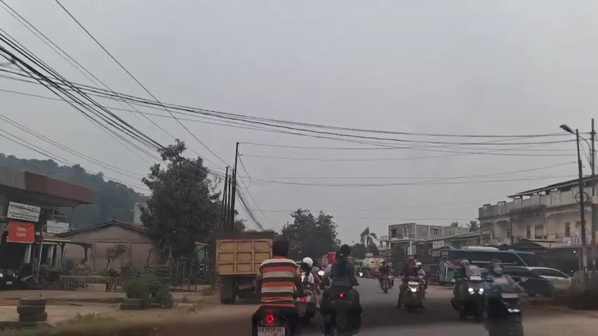 The video shows a street scene with multiple vehicles, including motorcycles, trucks, and a bus. The sky is overcast, and the environment appears to be somewhat rural or suburban, with buildings and trees visible along the roadside. Power lines are strung across the sky, and there is a truck with a yellow bed in the foreground, partially obscuring the view of a person on a motorcycle behind it. Traffic appears to be moving, though there might be some congestion. The overall impression is a typical day on a busy road in a populated area.