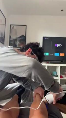 A man in a gray, long-sleeved shirt uses an electrical massage device on the lower leg of another person who is lying on a bed. The man using the device has dark curly hair, glasses, and black socks on his feet. The device is white with a blue light coming from the bottom. The person on the bed is obscured from the waist up. In the background, a television is on, displaying what appears to be a streaming service, and behind that is a white shelving unit with items in it. A framed landscape picture hangs on the wall to the left, along with a lamp, and another framed picture is partially visible.
