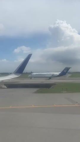The video shows a Lao Airlines plane on a runway, seen from the window of another aircraft. The tail and wing of the plane from which the video is shot are visible. The Lao Airlines plane is white with blue markings and a flower on its tail. The runway is gray and surrounded by grass. The sky is cloudy with patches of blue.