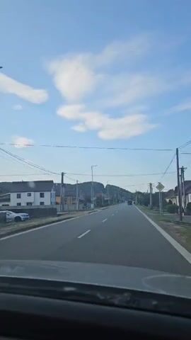 The video is an interior shot from a vehicle, looking forward through the windshield. The vehicle is moving along a paved road. On the left side of the road are houses and a sidewalk. On the right side of the road is a similar residential area. The road is a two-lane road with a white dashed line separating the lanes. There are power lines running along the sides of the road, supported by utility poles. The sky is blue with scattered white clouds. In the distance, there are hills. The time stamp indicates the video is a short clip.