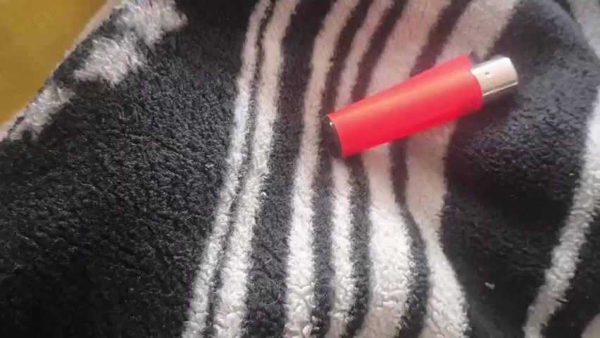A close-up shot shows a red lighter with a silver tip resting on a black and white striped surface. The lighter is positioned diagonally across the frame. The background appears to be a textured fabric.