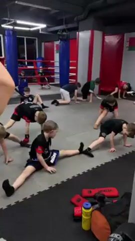 A group of approximately ten children are in a brightly lit indoor space that appears to be a martial arts training facility or gym. Most of the children are on the floor, performing stretching exercises. Some are in a full split, while others are in a modified stretch with one leg extended to the side. They are wearing a mix of athletic attire, including shorts and t-shirts, with some having visible logos. In the background, there is a boxing ring with blue corner posts, as well as red padded wall sections. Additional training equipment, such as punching bags and pads, are scattered around the room. There is a person partially visible in the lower left corner, presumably an instructor or observer. The floor is covered in gray interlocking foam mats. Overall, the setting suggests a structured training session focused on flexibility.