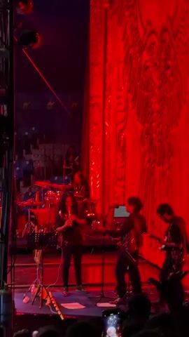The video opens on a stage where a band is performing. Red lights dominate the scene. In the foreground, a person is playing a wind instrument. To their right, two other individuals are playing guitars. Behind them, a drummer is seated at a drum kit. The stage has a tall metal structure on the left side, and a large, elaborate red curtain or backdrop on the right. The audience is visible in the background, seated in tiered rows.

The scene then transitions to a performance involving motorcycles inside a large, cylindrical structure. The structure is made of metal lattice and illuminated by red lights. Several dirt bikes are visible, some with riders. The riders appear to be wearing protective gear, including helmets. The motorcycles are moving within the cylinder, suggesting a stunt performance. The audience is again visible, watching the performance. The lighting remains predominantly red, creating a dramatic and intense atmosphere. The video captures a dynamic and exciting show, likely a concert with a motorcycle stunt element.