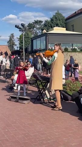 The video captures an outdoor musical performance in Petrovaradin, Serbia, on a clear, sunny day. In the foreground, an adult woman and a young girl are playing violins. The woman, wearing a long beige dress over a patterned top, stands to the right, while the girl, dressed in a red coat and white tights, stands to the left, both focused on their instruments. Behind them, a man wearing sunglasses and a white top plays a keyboard set up on a stand. Two violin cases are open on the reddish brick ground near the performers, suggesting a street performance or busking.

Numerous people are visible in the background, some seated on benches, others standing or walking by, observing the performance or enjoying the sunny weather. A modern-looking building with large glass windows and an older structure are seen behind the crowd, surrounded by trees and shrubs. A street light pole with multiple lamps is also present. The overall scene depicts a lively and pleasant atmosphere.